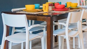 A dining area with a dark blue wall, a wooden geometric shelf with plants, and a table set with colorful bowls and white chairs.