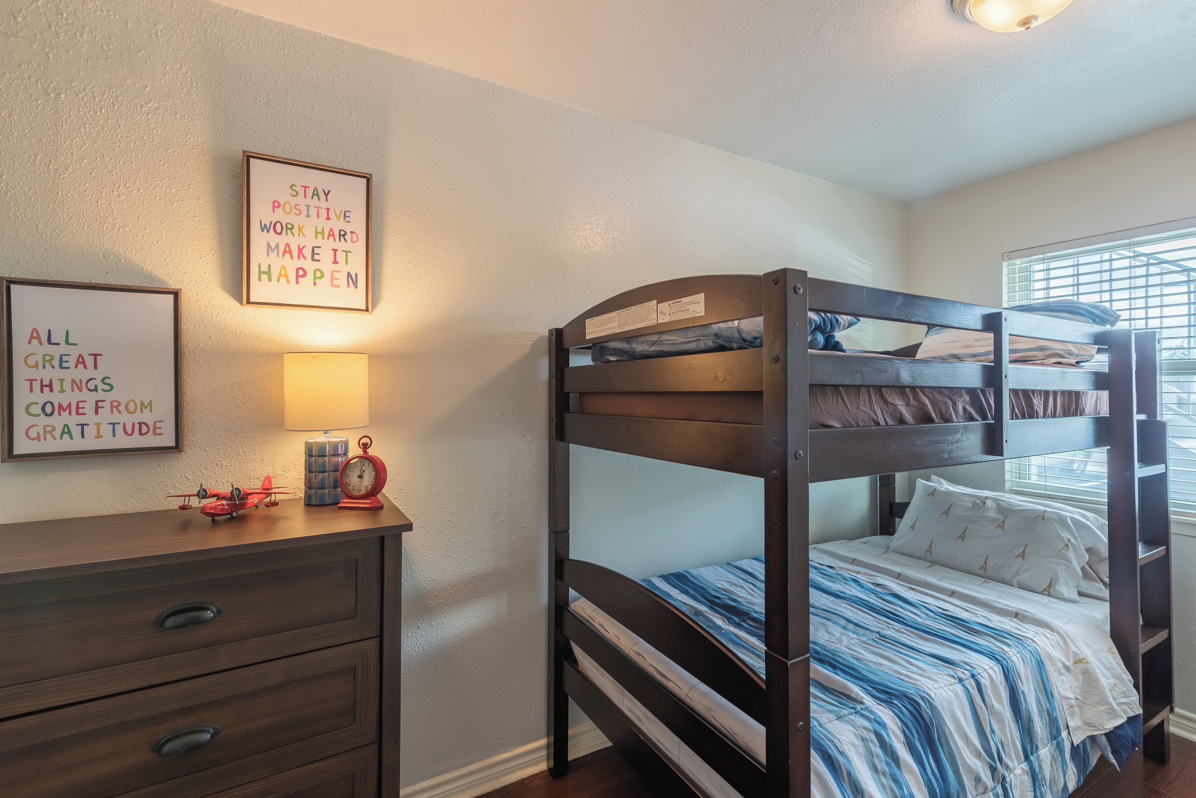 Un lit superposé avec une couvre-lit à rayures, une commode avec des tiroirs, une lampe et des affiches motivantes accrochées au mur.
