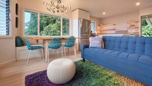 Een moderne keuken met een inbouwkoelkast, een eethoek met een tafel en stoelen, en een blauwe bank met een kussen waarop 'ALOHA' staat.