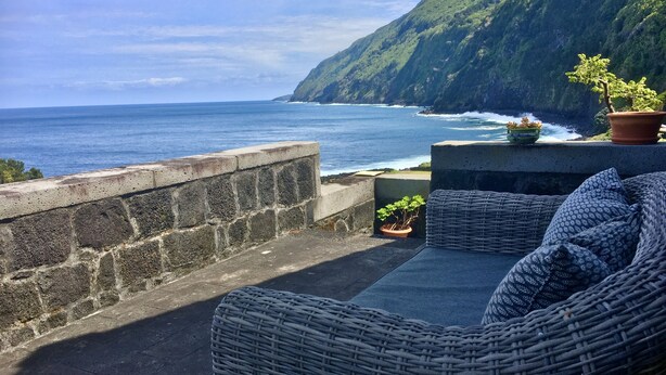 A wicker sofa with cushions, a potted plant, and a view of the ocean and cliffs.