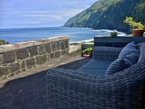 A wicker sofa with cushions, a potted plant, and a view of the ocean and cliffs. - Enchanting view from the balcony of beautifully restored "Adega Baia do Canto". (Santo Amaro, Sao Roque, Ilha do Pico, Azores)