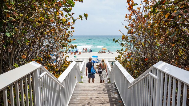 Eine Holztreppe führt zu einem Strand mit Menschen und Sonnenschirmen.