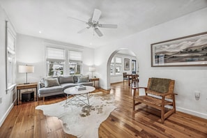 A spacious living room with hardwood floors, a ceiling fan, a sofa, a coffee table, and a rug. - Historic Beach Cottage - Steps to Beach & Dining (Saint Simons Island)