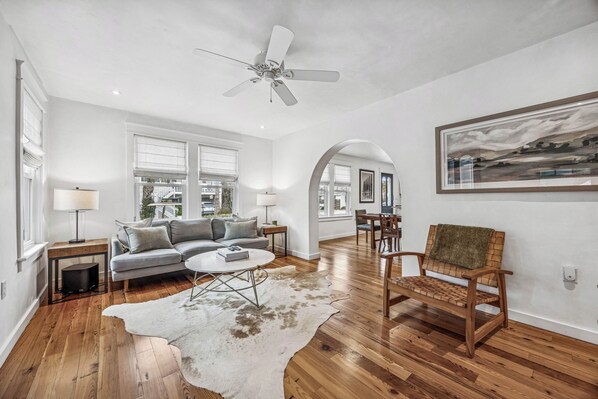 A spacious living room with hardwood floors, a ceiling fan, a sofa, a coffee table, and a rug. - Historic Beach Cottage - Steps to Beach & Dining (Saint Simons Island)