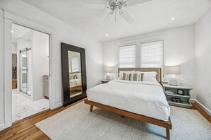 A bedroom with a wooden bed, white bedding, a large mirror, bedside lamps, and a ceiling fan. - Historic Beach Cottage - Steps to Beach & Dining (Saint Simons Island)