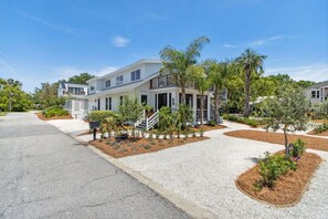 A modern two-story house with a front porch, a driveway, and landscaped gardens with palm trees. - Historic Beach Cottage - Steps to Beach & Dining (Saint Simons Island)