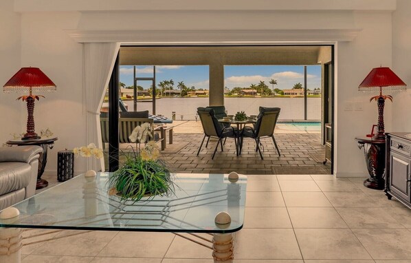 A modern living room with a glass coffee table, a sofa, and two lamps. There is a view of an outdoor patio with a pool and chairs.