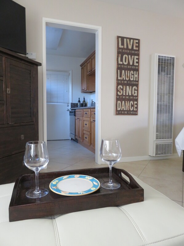Un comedor con una bandeja que sostiene dos copas de vino y un plato, una cocina visible a través de una puerta abierta, y una cita motivacional en la pared.