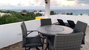 Ett takförsett utomhusområde med sittplatser, bestående av flätade stolar och ett runt bord, med utsikt över kusten, bebyggelsen och havet.