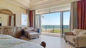 A spacious bedroom with a large bed, two armchairs, a dresser, and a view of the ocean through large sliding doors.