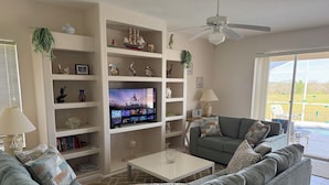 A living room with a flat-screen TV, grey sofas, a coffee table, and a bookshelf filled with decorative items.