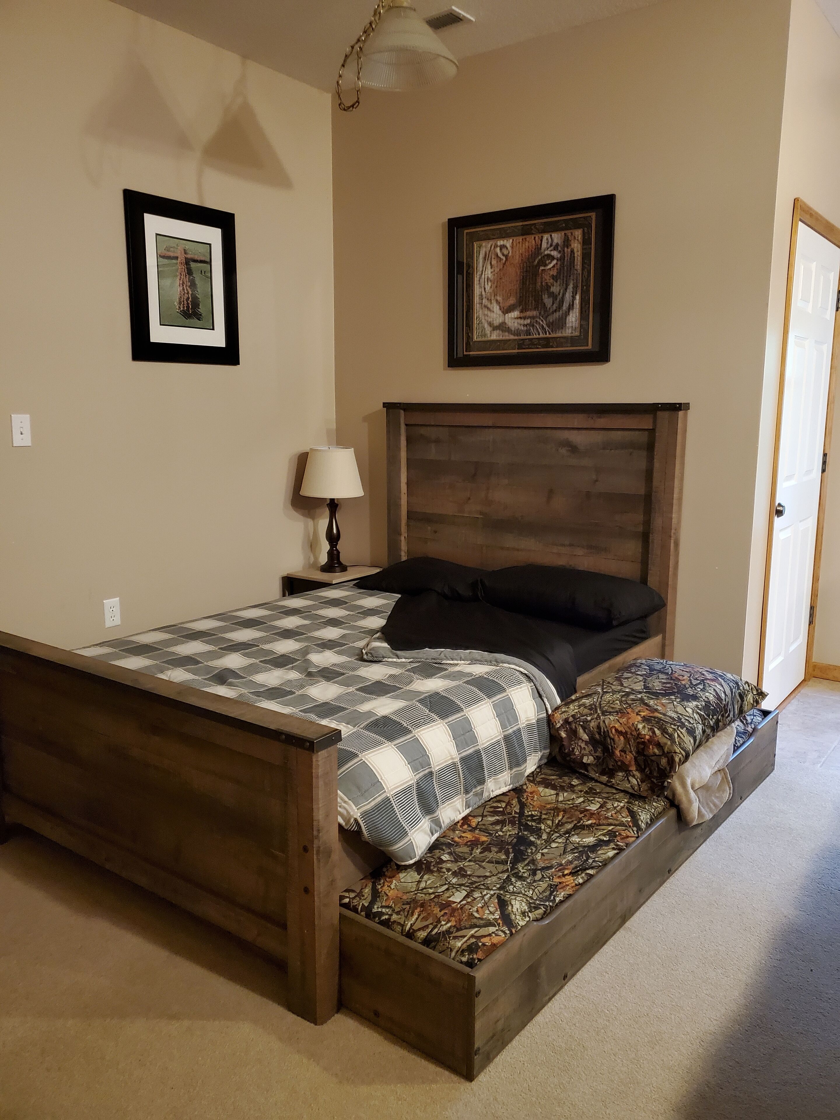 Une chambre à coucher avec un cadre de lit en bois, une couvre-lit à carreaux, un coussin à motifs, une table de chevet avec une lampe et deux tableaux encadrés accrochés au mur.