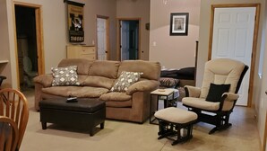 A living room with a brown sofa, a black ottoman, a beige armchair, and a wooden chair. There is a ceiling fan, a framed picture on the wall, and a door in the background. - Sander Ridge - 40 wooded acres (Columbia)