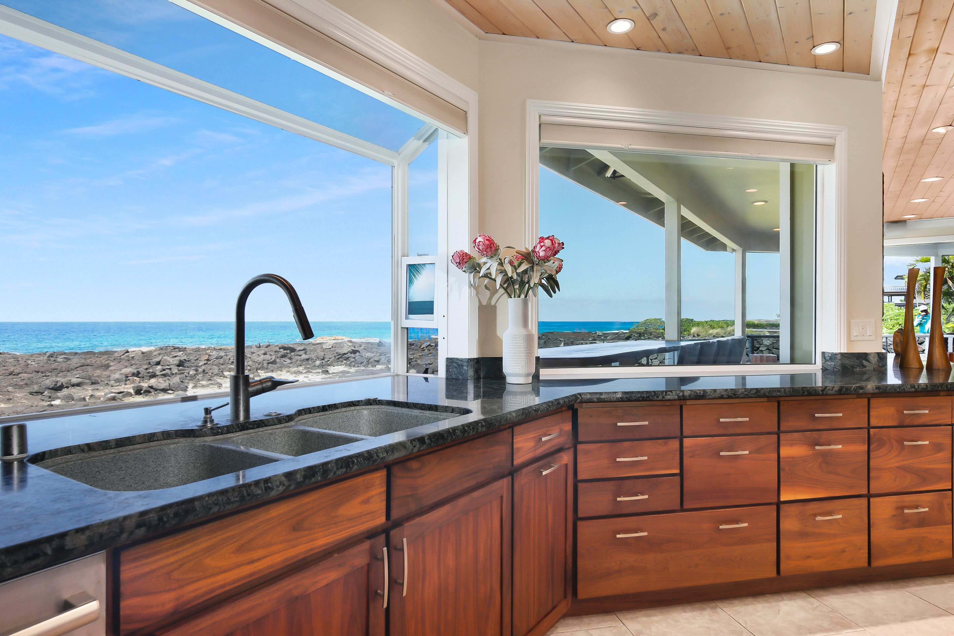 A kitchen with a large window overlooking a beach, wooden cabinets, and a granite countertop.