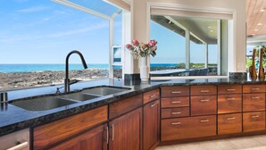 Una cocina con un amplio ventanal con vistas a una playa, muebles de madera y encimera de granito.