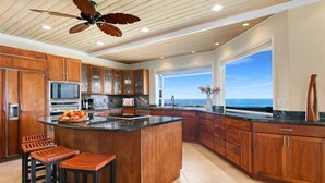 Una cocina amplia con armarios de madera, una isla grande con taburetes, un ventilador de techo y vista al océano a través de un ventanal.