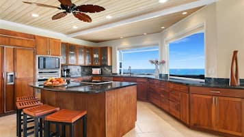 A spacious kitchen with wooden cabinets, a large island with stools, a ceiling fan, and a view of the ocean through a large window.