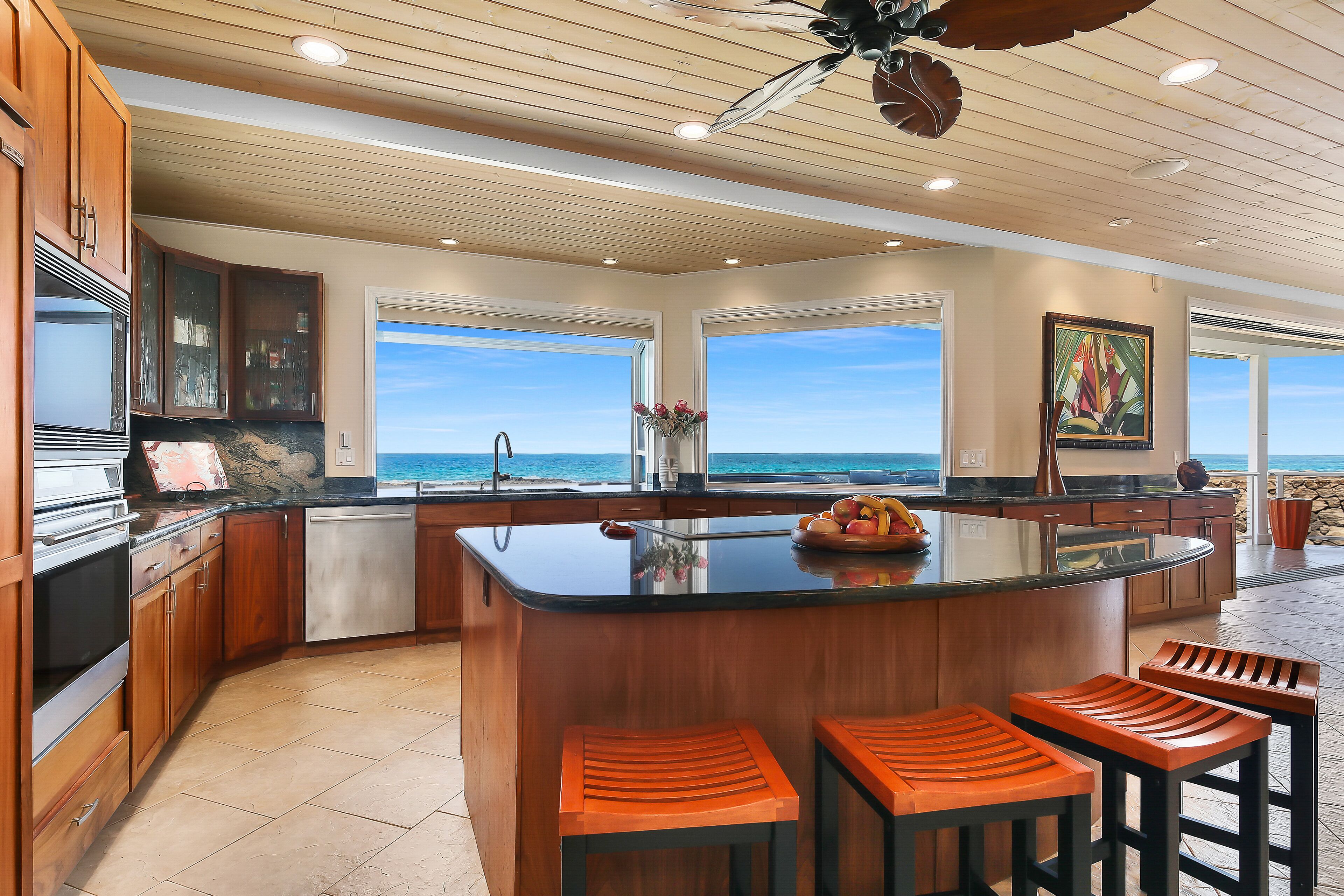 A modern kitchen with wooden cabinets, a central island with bar stools, and large windows overlooking the ocean.