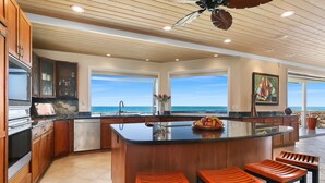Una cocina moderna con armarios de madera, una isla central con taburetes altos y amplias ventanas con vistas al océano.