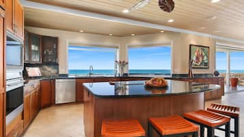A modern kitchen with wooden cabinets, a central island with bar stools, and large windows overlooking the ocean.