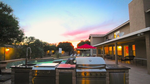 Et udendørs køkkenområde med grill og jacuzzi, omgivet af en pool og patio møbler.