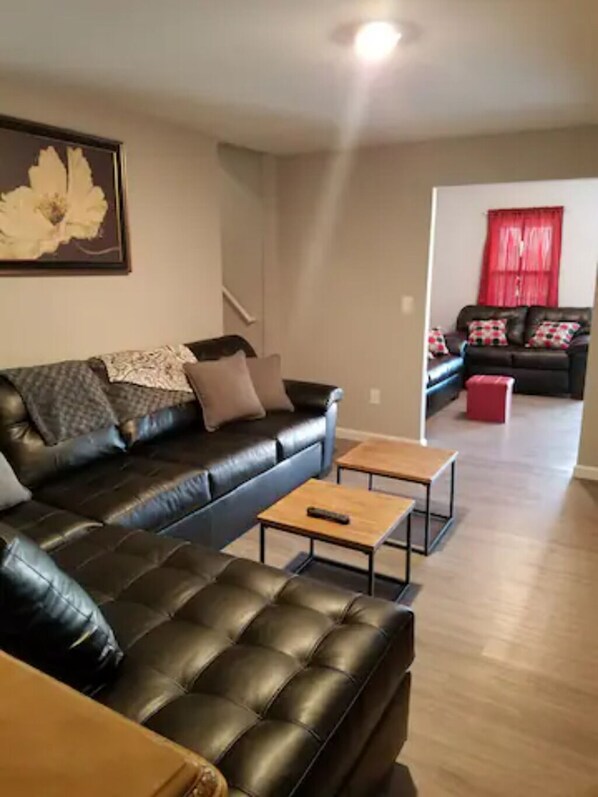 A living room with a black leather sofa, a wooden coffee table, and a red ottoman. There is a framed artwork on the wall and a staircase leading to another room. - The Carter's Wildwood Rentals- Master Suite #3 (Wildwood)