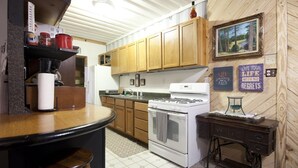 A kitchen with wooden cabinets, a stove, and a sink. There are various kitchen items on the counter, and a painting on the wall. - Private 50 ft waterfall. Eco-friendly, cozy shipping container cabin (Tracy City)