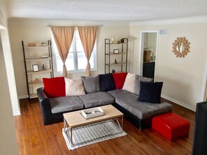 A modern living room with a grey sectional sofa, red and black cushions, a wooden coffee table, and a red ottoman.