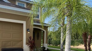 A residential house with a garage, a driveway, and a landscaped garden with palm trees. - Amazing 5 bedroom house near Disney (Kissimmee)