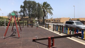 A playground with swings and climbing structures, a parking area with a car, and a fence with colorful vertical stripes. - Beautiful villa with heated pool, large patio area and barbecue (Fuerteventura)
