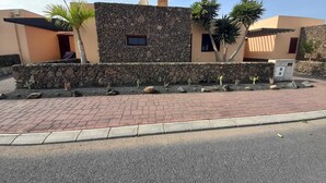A modern building with a stone facade, a paved driveway, and a small garden with cacti and palm trees. - Beautiful villa with heated pool, large patio area and barbecue (Fuerteventura)