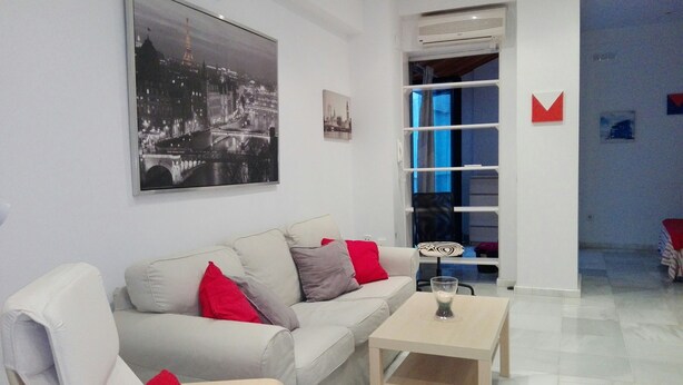 Een moderne woonkamer met een witte bank, rode kussens, een klein houten bijzettafeltje en een stadsgezichtschilderij aan de muur.