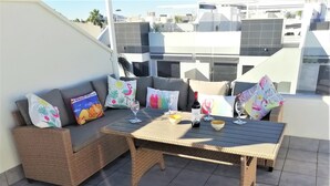 Un balcón con sofá de mimbre, una mesa con copas de vino y cojines decorativos.