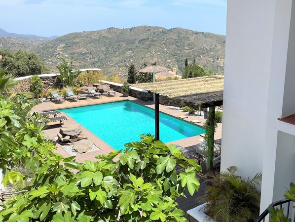 A rectangular swimming pool with lounge chairs and a stone wall, surrounded by greenery and a mountainous backdrop.