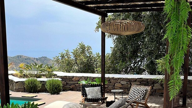Un área de estar al aire libre con techo, piscina, paredes de piedra y vistas a las colinas.