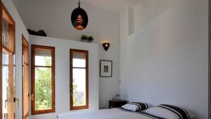 Un dormitorio con una cama grande, dos ventanas, una lámpara colgante y una mesita de noche con lámpara.