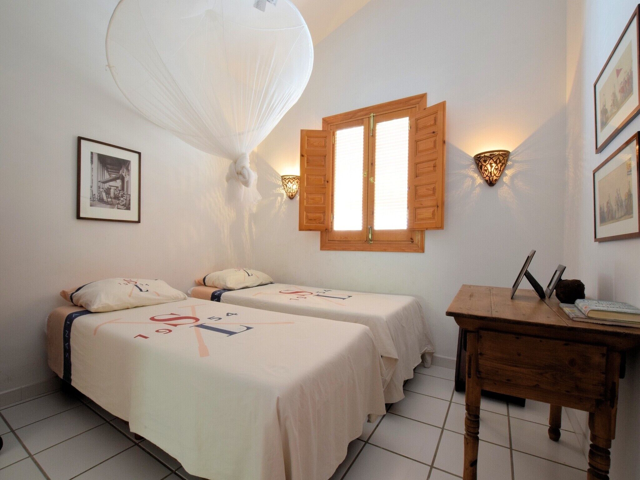 A hotel room with two beds, a wooden desk, a wall-mounted lamp, and a window with shutters.