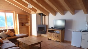 A cozy living room with a wooden ceiling, a fireplace, a television, and a small kitchen area.
