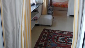 Un couloir étroit avec un tapis à motifs, un lit recouvert d’une courtepointe colorée et une fenêtre avec des rideaux.