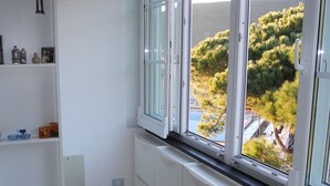 Une pièce d’angle avec une fenêtre donnant sur une colline, des murs blancs et une étagère où sont disposés divers objets.