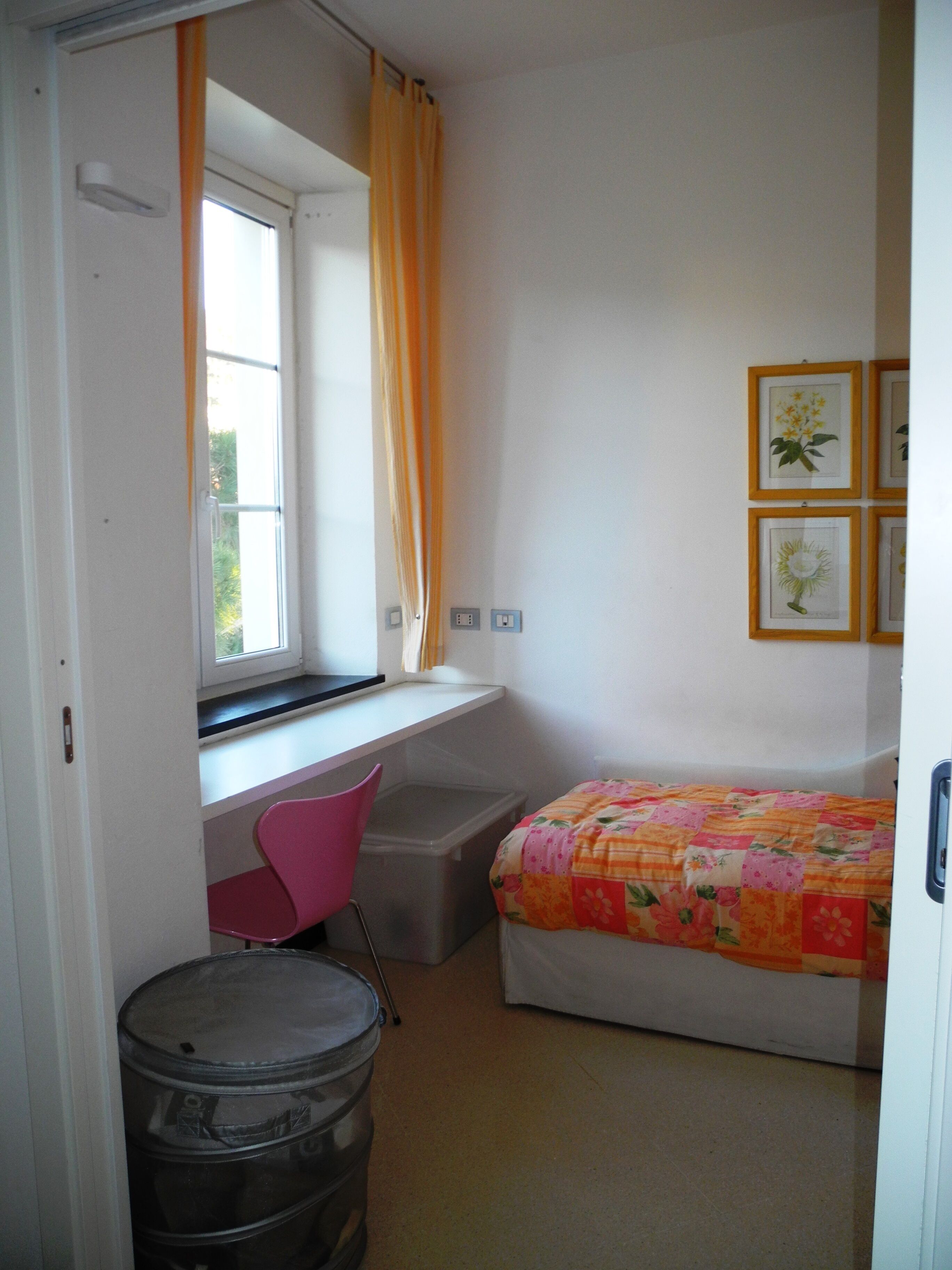A bedroom with a bed, a desk, a chair, a window with curtains, and framed pictures on the wall.
