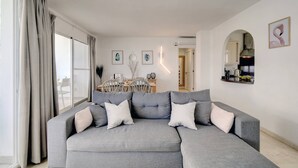 A living room with a grey sectional sofa, a dining area with a table and chairs, and a kitchen visible through an archway. - Astor Estepona: Lux 2BD Frontline Marina, Pool, WiFi, Sea views, Private parking (Estepona)