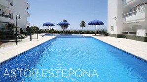 A swimming pool with blue water, surrounded by white buildings and blue umbrellas. - Astor Estepona: Lux 2BD Frontline Marina, Pool, WiFi, Sea views, Private parking (Estepona)