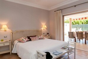 Une chambre à coucher comprenant un lit, des tables de chevet, une commode, un présentoir à livres et donnant sur un espace extérieur aménagé pour les repas.