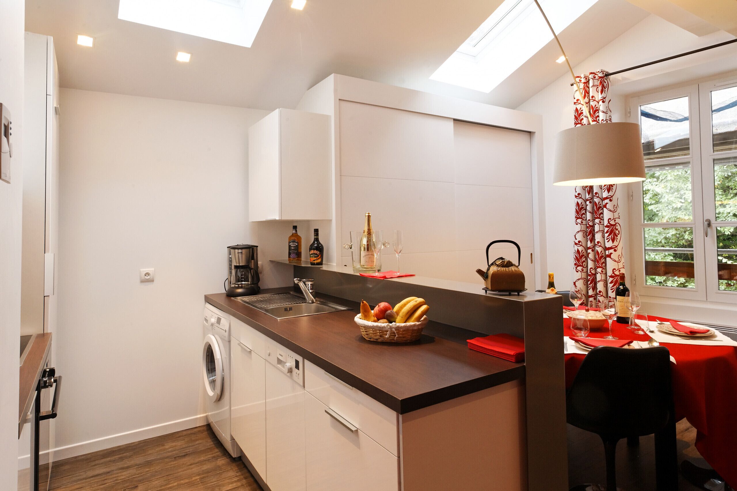 A modern kitchen with a built-in washing machine, a dining area with a red tablecloth, and a window with a view of greenery.