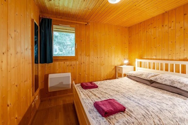 Une chambre dans un chalet en bois, avec un lit, une table de chevet, une fenêtre avec des rideaux et un radiateur.