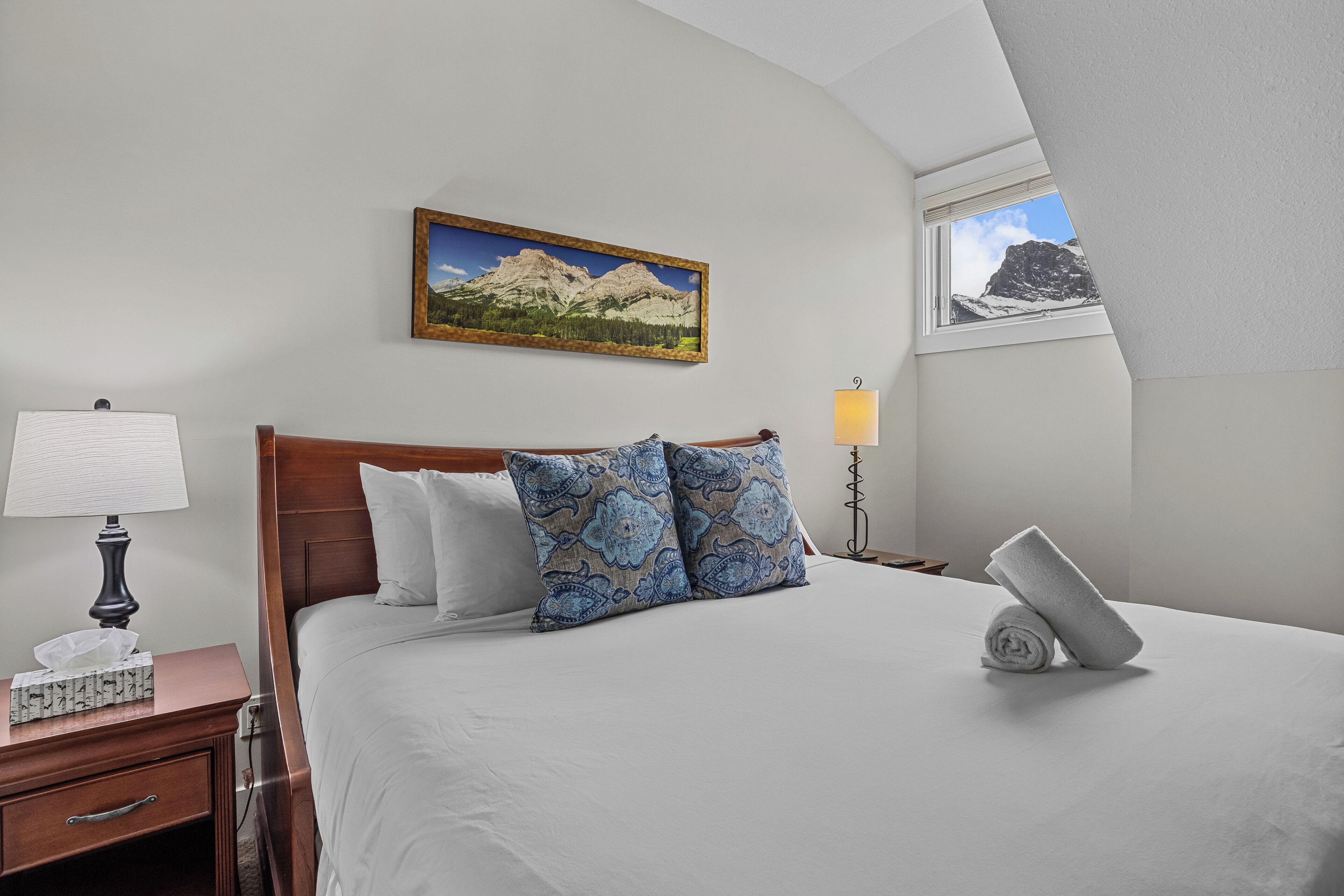 A neatly made bed with a mountain landscape painting above it, a bedside lamp, and a window with a mountain view.