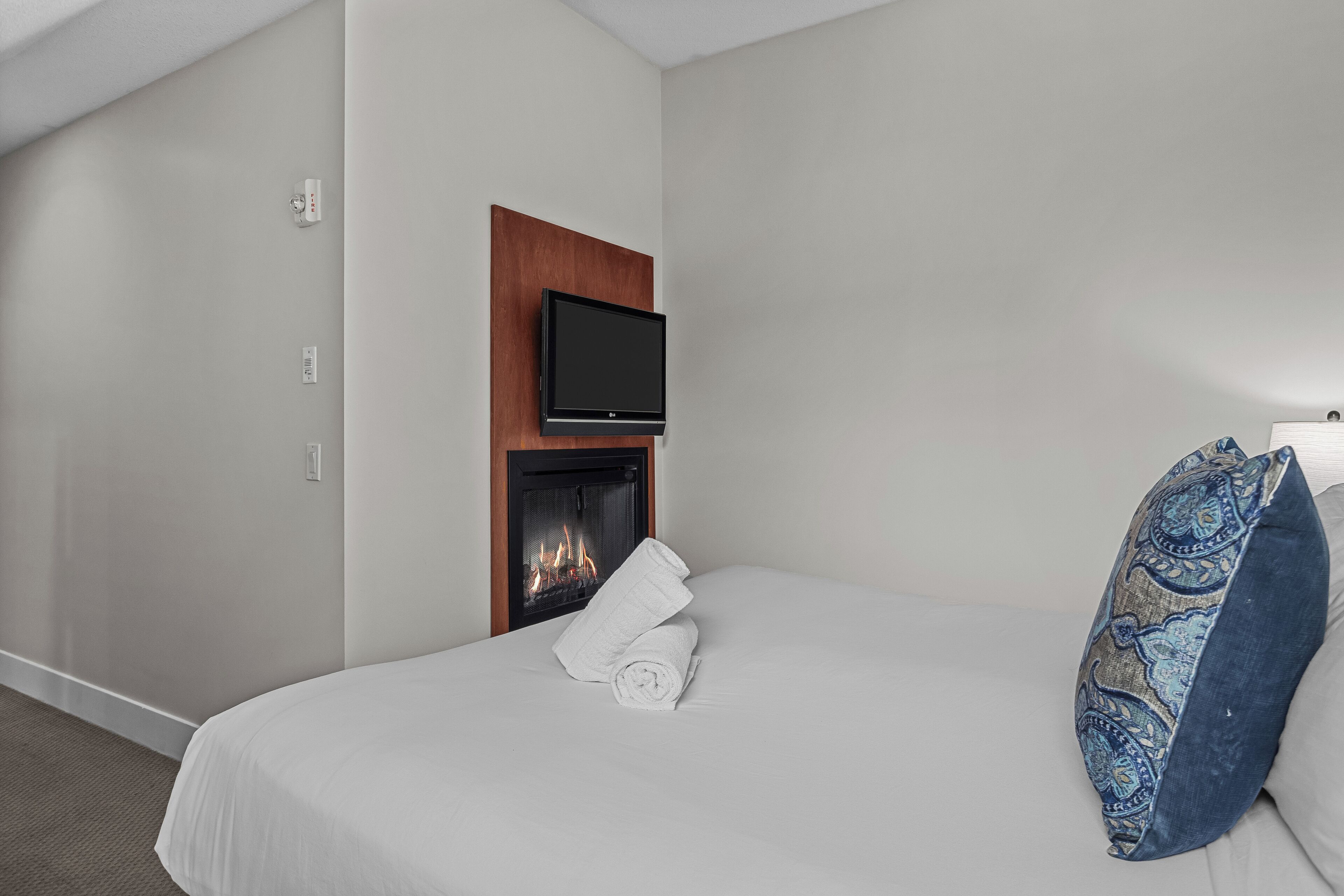A hotel room with a bed, a flat-screen TV mounted on a wooden panel above a fireplace, and a pair of rolled towels on the bed.