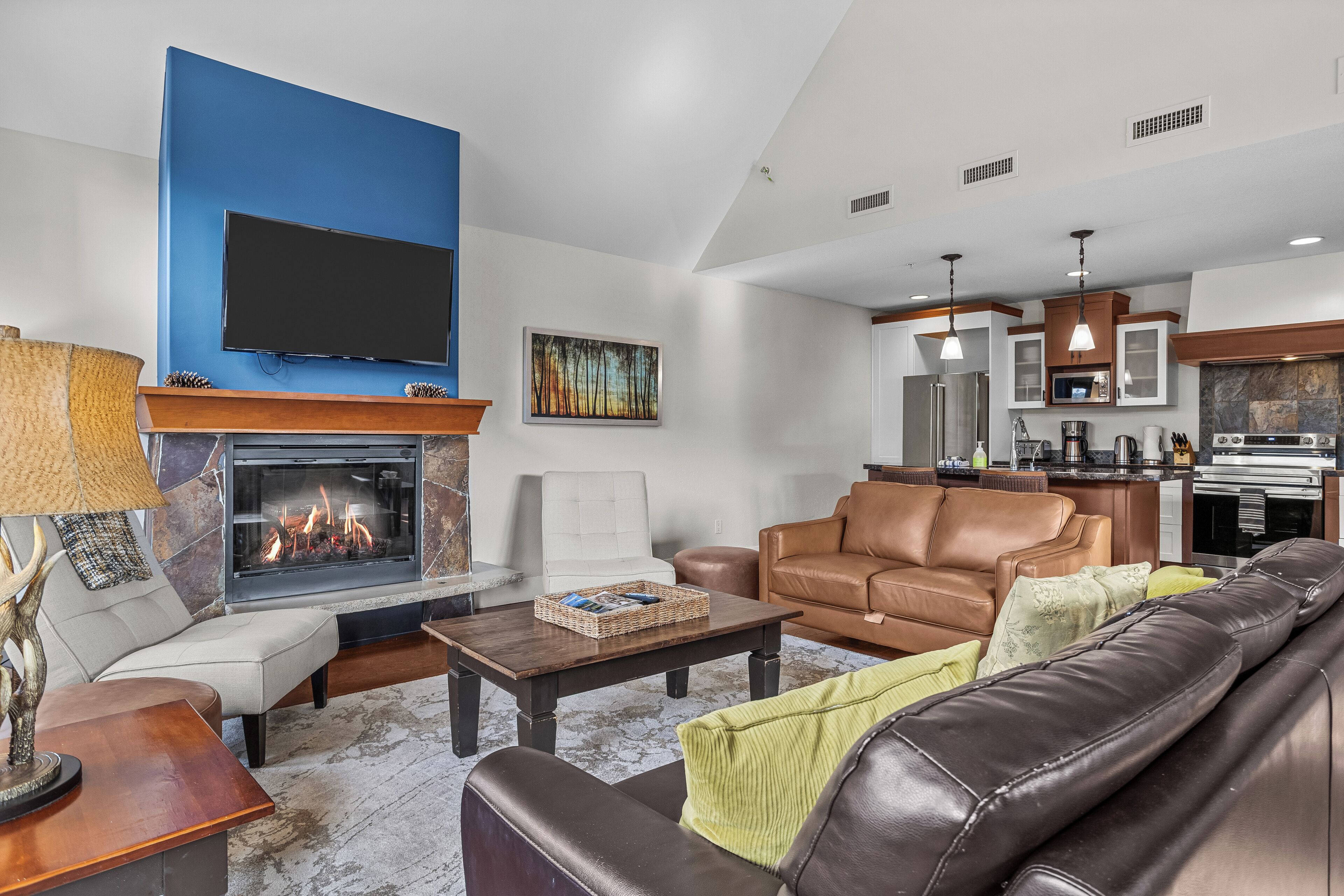 A living room with a fireplace, leather sofas, a flat-screen TV, and a kitchen area.