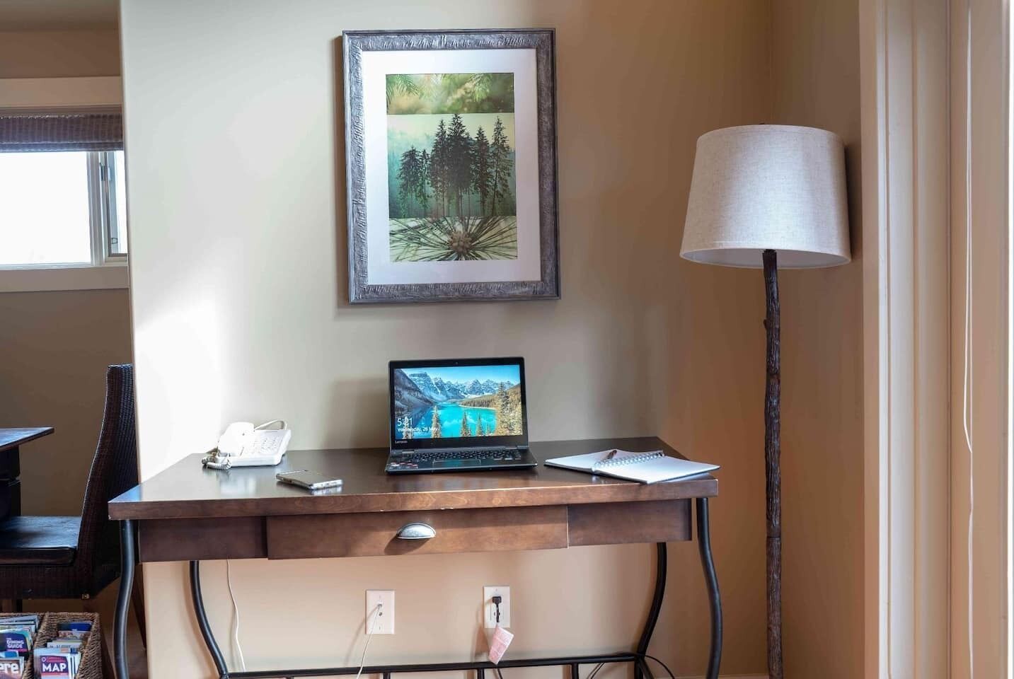 A hotel room with a wooden desk, a laptop, a lamp, and a framed picture on the wall.